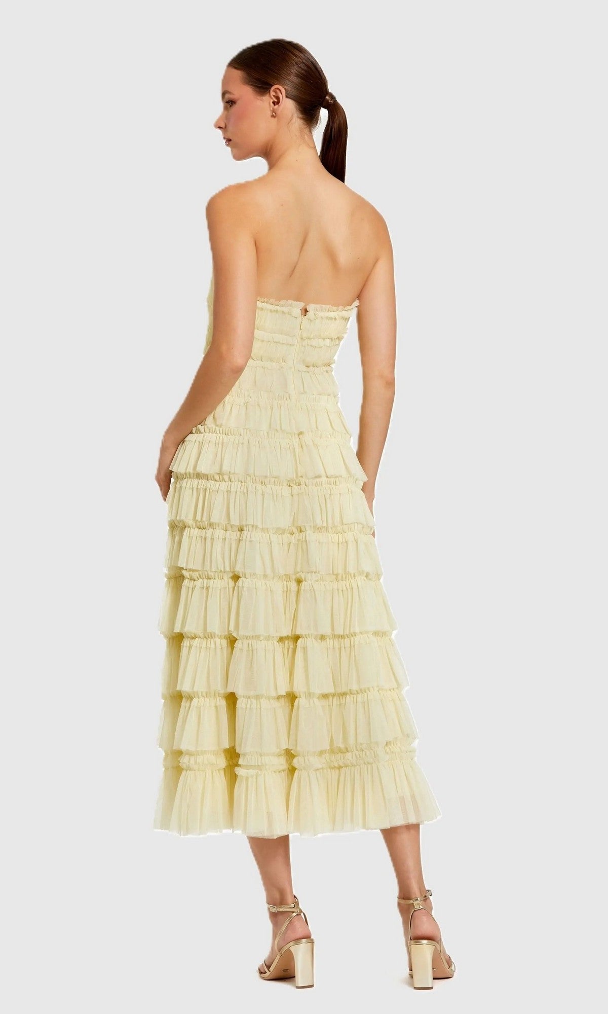 A woman stands with her back to the camera in a Mac Duggal 8286 midi semi-formal dress featuring micro-ruffle detailing, paired with gold sandals, against a plain light gray background.